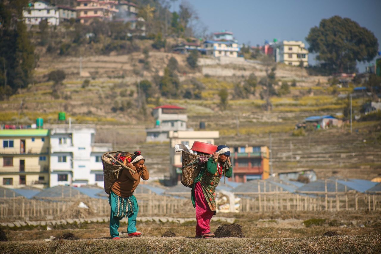 Two Nepalese women carrying baskets through a rural field against a village backdrop.